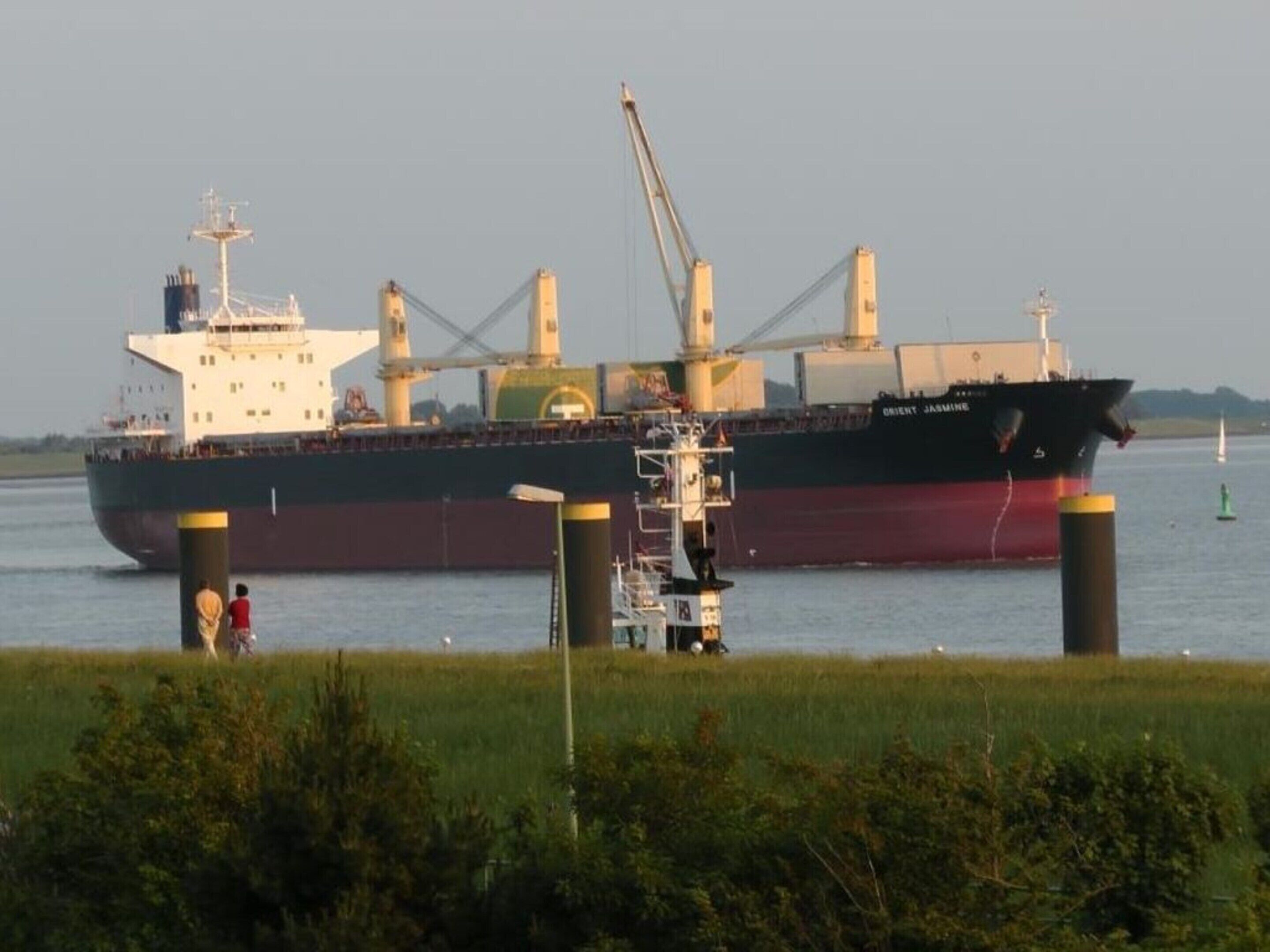 Una grande nave cargo ormeggiata in un porto, con gru e una piccola imbarcazione nelle vicinanze.