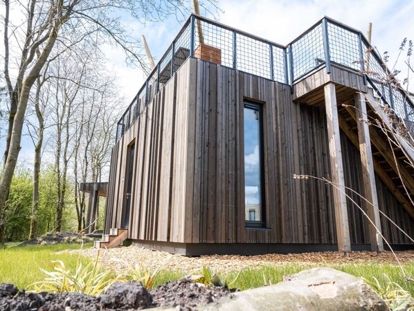 A modern wooden cabin with a balcony, surrounded by trees and a grassy area.