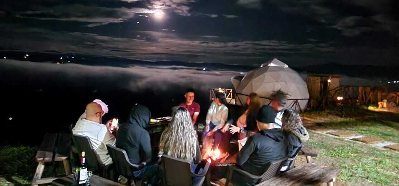 Un grupo de personas reunidas alrededor de una fogata por la noche, con una tienda de campaña y una estructura en forma de cúpula al fondo.