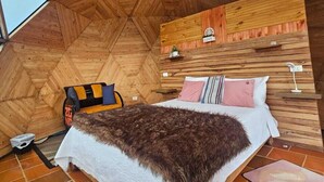 Una habitación con techos altos y suelo de madera en dibujo de espiga, una cama con manta blanca y marrón, una mesita y un estante montado en la pared con un cartel.