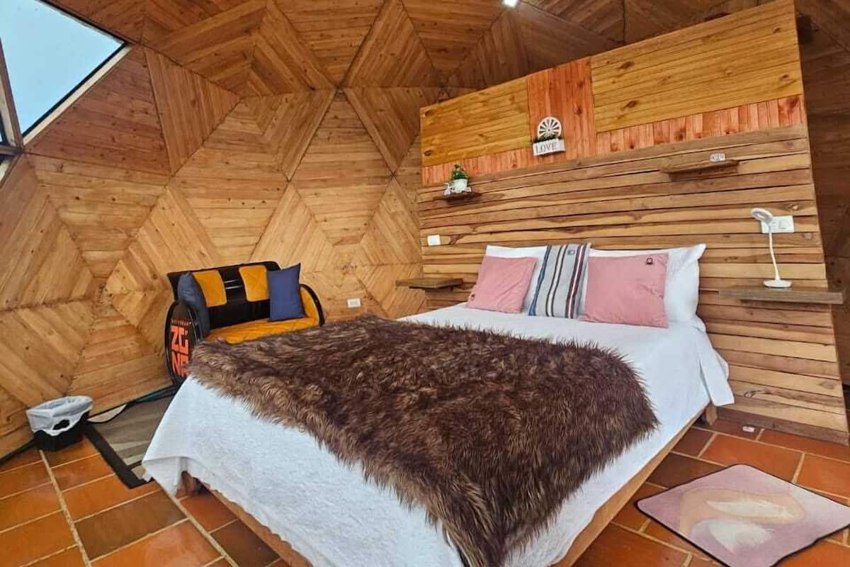 A high-ceilinged room with a wooden herringbone floor, a bed with a white and brown blanket, a small table, and a wall-mounted shelf with a sign.