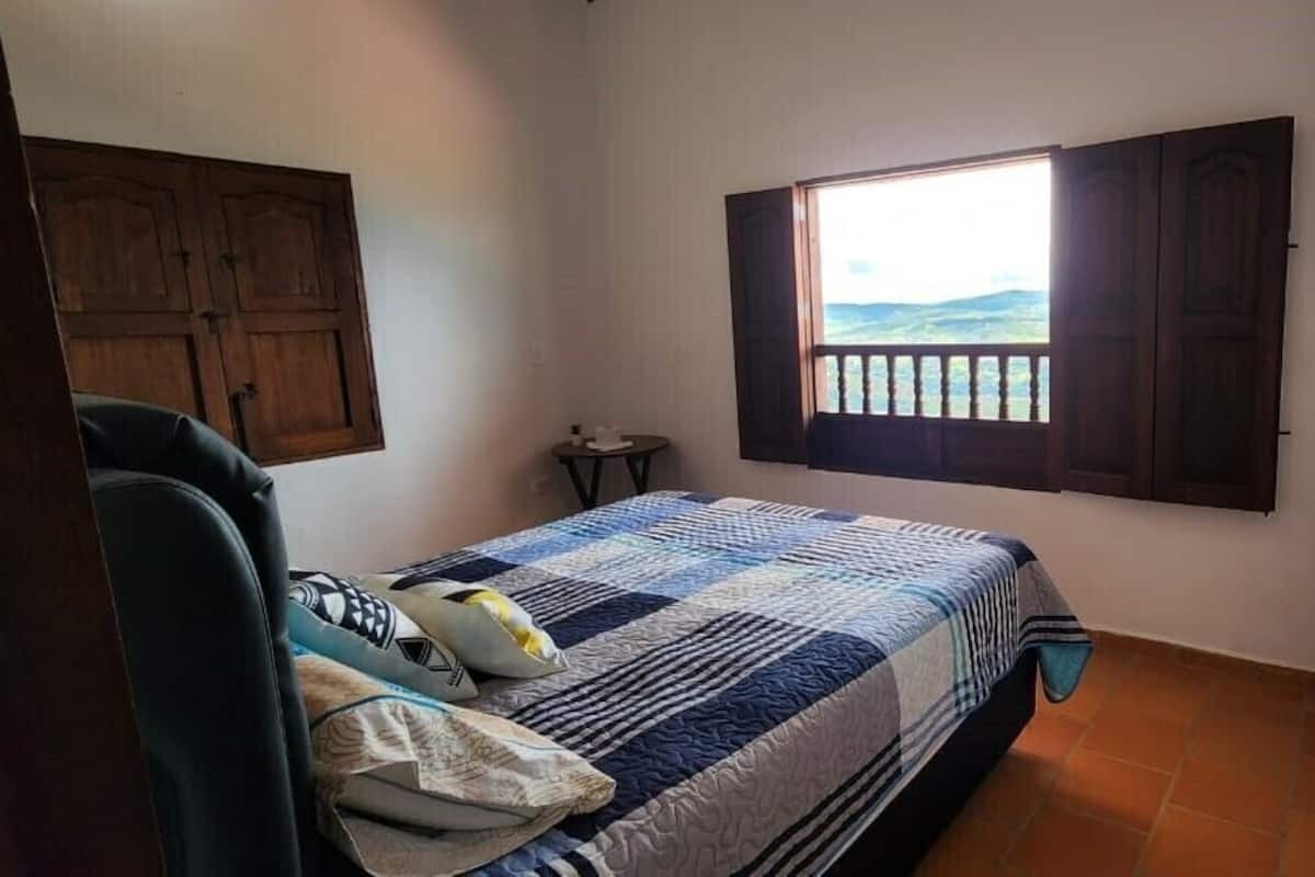 A bedroom with a bed, a window with shutters, a small table, and a chair.