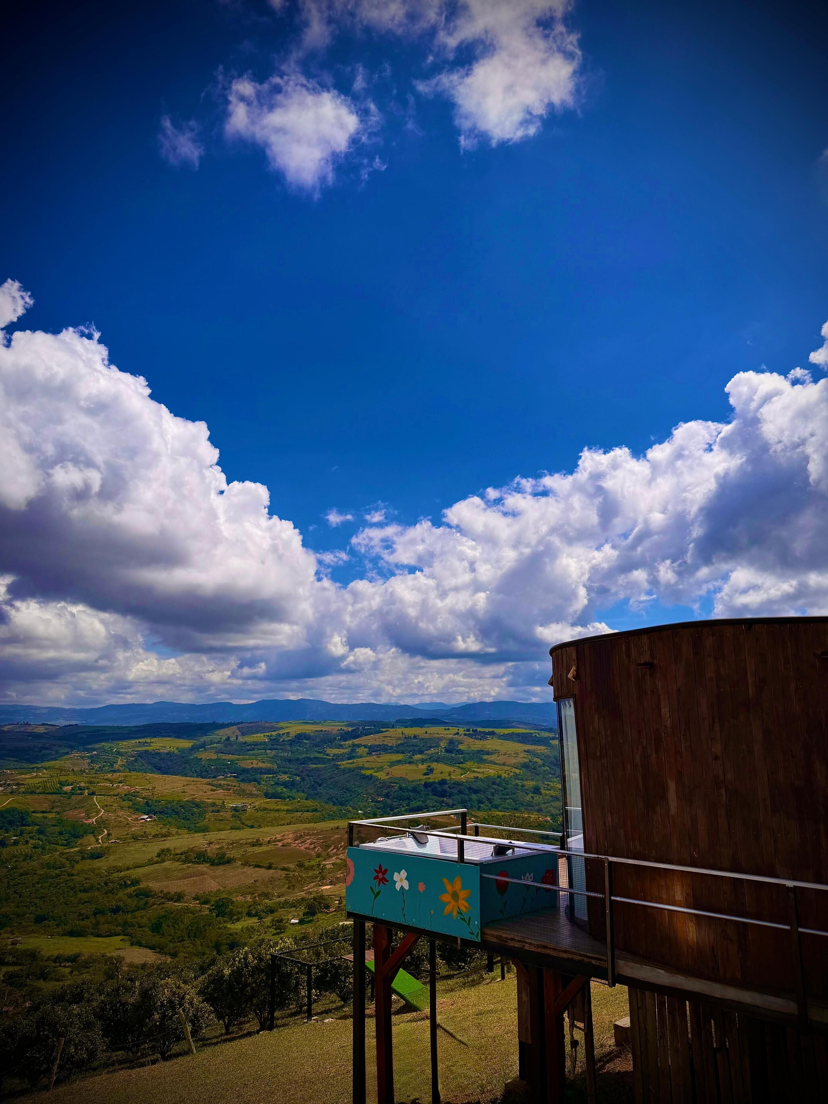 Una estructura de madera con vistas a un paisaje verde y montañas distantes bajo un cielo azul con nubes.