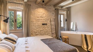 Un dormitorio con una cama grande, una pared de piedra, un ventanal con cortinas, una lámpara de noche y vistas al verdor exterior.