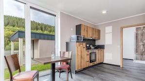 Una cocina moderna con armarios de madera, microondas y estufa. Hay una mesa de comedor con sillas y vistas a un bosque a través de amplios ventanales.