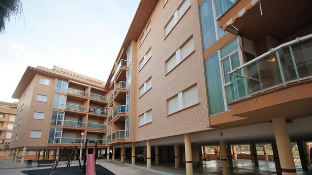 Un edificio residencial de varios pisos con balcones, estacionamiento techado y un área de juegos.