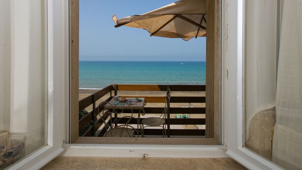 Uitzicht vanuit een kamer met een houten bankje en tafel, op een strand met heldere lucht en een kalme zee.