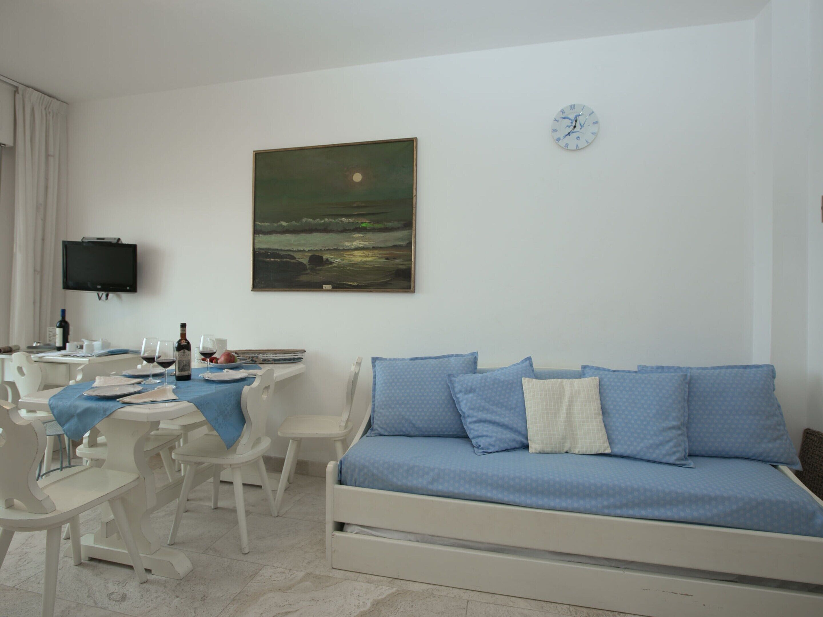 Een kamer met een bed, een tafel met stoelen, een schilderij, een klok en een televisie aan de muur.
