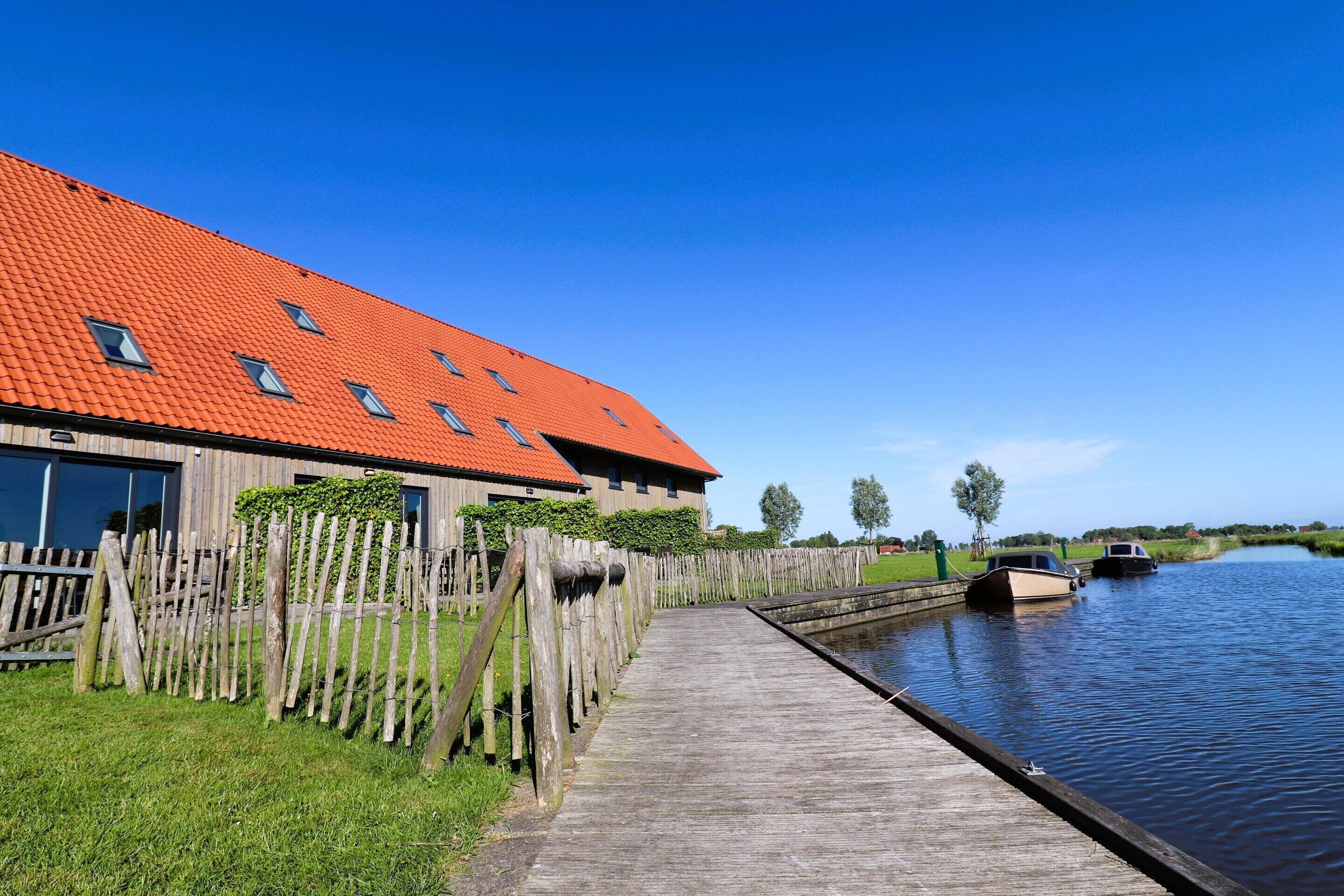 Un muelle de madera que conduce a un canal con un bote, un edificio de techo rojo y un cielo azul despejado.
