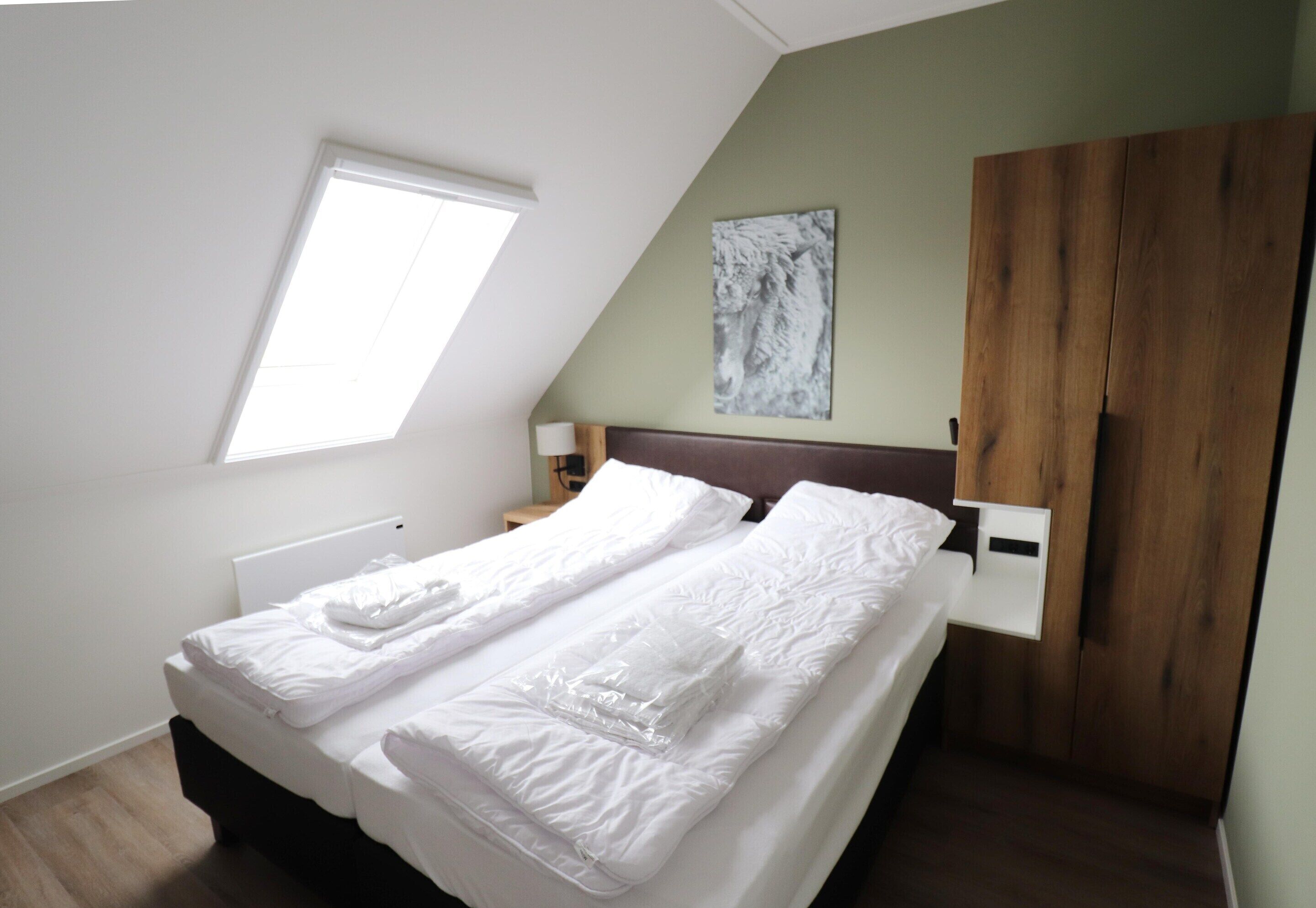 A bedroom with a bed, a window, a wooden wardrobe, and a painting on the wall.