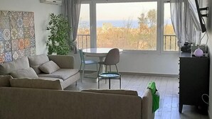 A living room with a sofa, a dining table with chairs, a potted plant, and a view of the city through large windows. - Belvilla by OYO Casa El Pinar (Torremolinos)