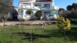 A two-story house with a light green facade, a red-tiled roof, and a small front yard with a tree and a bush. - Apartments Kozino - Two Bedroom Apartment (Zadar)