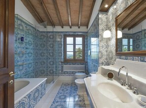 A bathroom with a large bathtub, a double sink, and a toilet. The walls are covered in blue patterned tiles, and there is a window above the toilet. - Villa Miniato Comfortable Holiday Residence (San Miniato)