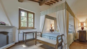 A bedroom with a four-poster bed, a wooden bench, a fireplace, and a window with a view of greenery. - Villa Miniato Comfortable Holiday Residence (San Miniato)