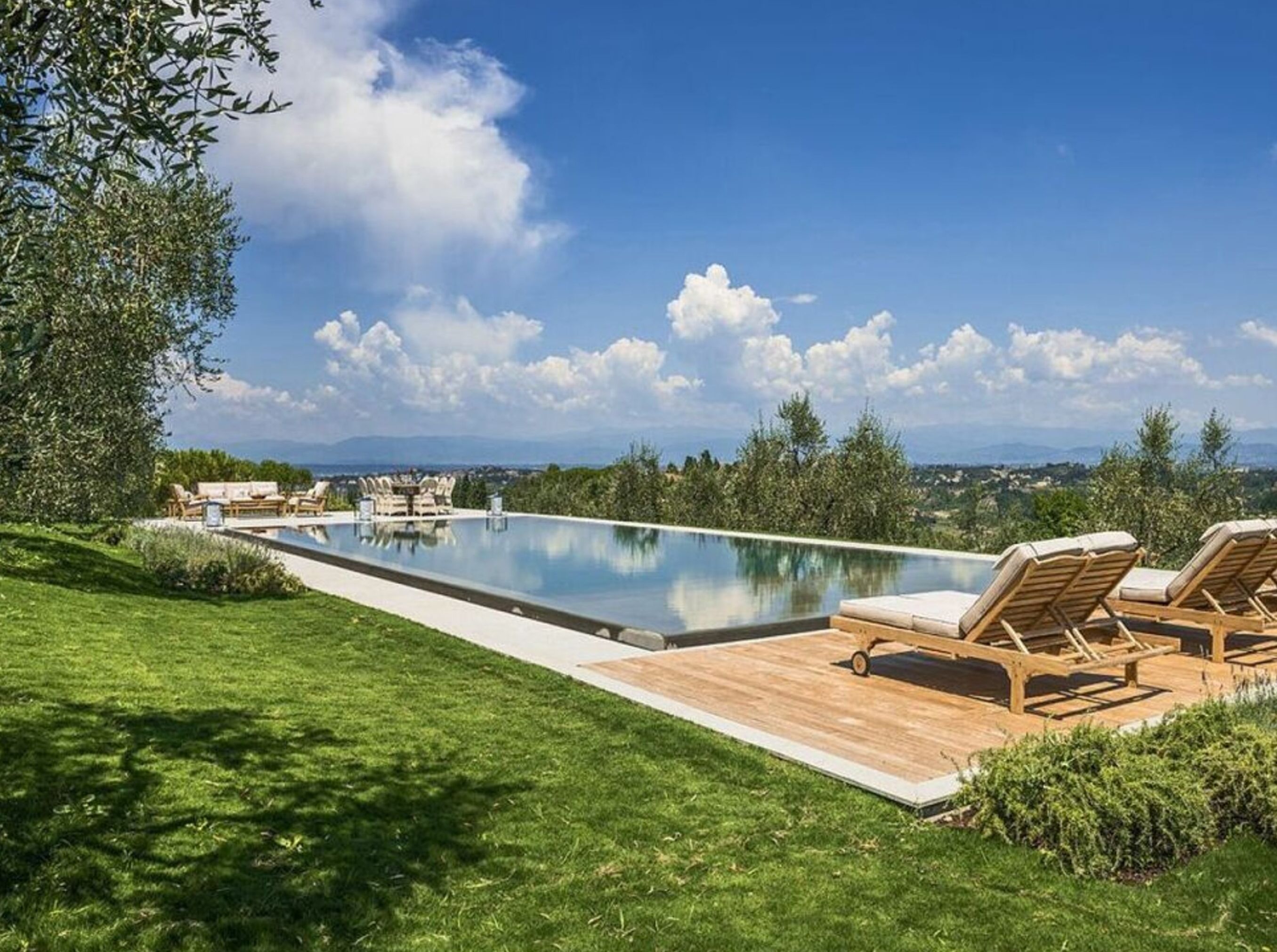 A swimming pool with lounge chairs and a wooden deck, surrounded by greenery and a clear sky.