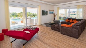 A spacious living room with a red sofa, a brown sofa with orange cushions, and a red ottoman. There is a large sliding glass door leading to an outdoor area with a pool and lounge chairs. - New White Villa, 60 m2, Pool (Vodnjan)