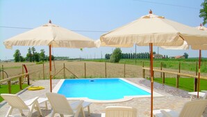 A swimming pool with white lounge chairs and beige umbrellas, surrounded by a stone patio. - Belvilla by OYO Casa Rosolina Due (Ariano nel Polesine)