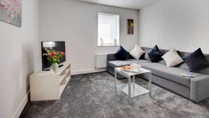 A modern living room with a grey sofa, a coffee table, a TV, and a vase of flowers. - Seasalt Twobedroom Apt in Skinningrove (Saltburn-by-the-Sea)