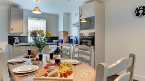 A modern kitchen with white cabinets, a built-in oven, and a microwave. A dining table with chairs is set with plates, a bottle, and a fruit platter. - Seasalt Twobedroom Apt in Skinningrove (Saltburn-by-the-Sea)