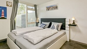 A bedroom with a bed, a nightstand, a lamp, a window with curtains, a framed picture, and a small shelf with a lamp. - Huijsmans Verhuur Type A de Luxe Grevelingen 29 (Ouddorp)