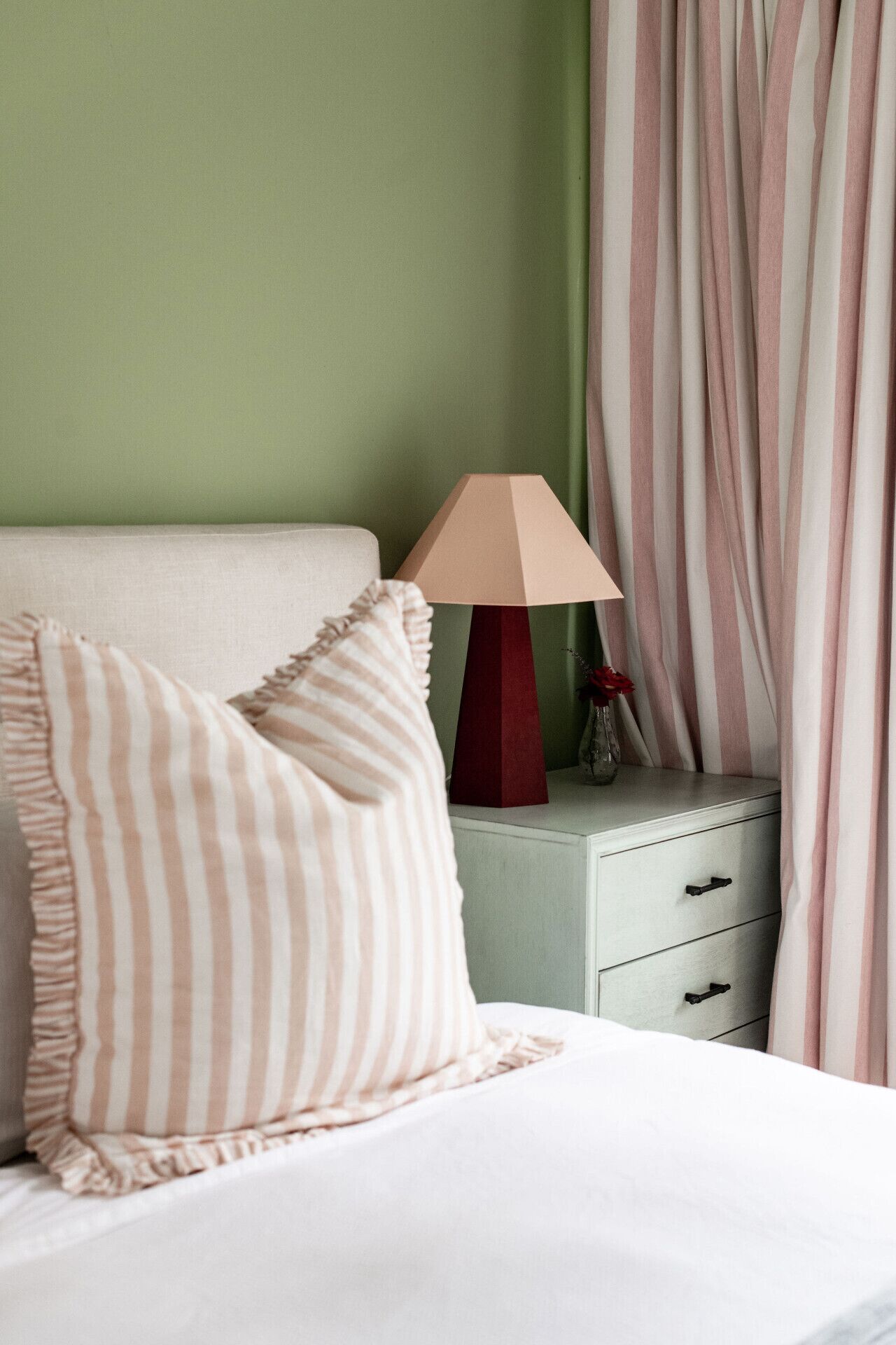 A bed with a striped pillow, a bedside table with a lamp, and a nightstand with a vase of flowers.