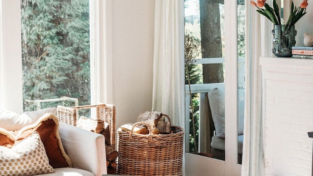 A cozy living room with a sofa, a wooden table with books, a wicker basket, and a view of greenery outside.