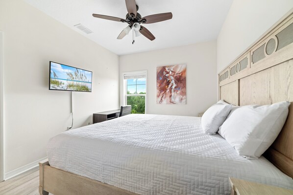 A bedroom with a bed, ceiling fan, TV, artwork, and a window.