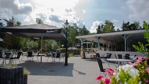An outdoor patio area with tables and chairs under umbrellas, a covered seating area, and a view of trees and sky. - Holiday Home With Covered Terrace in Rhenen (Rhenen)