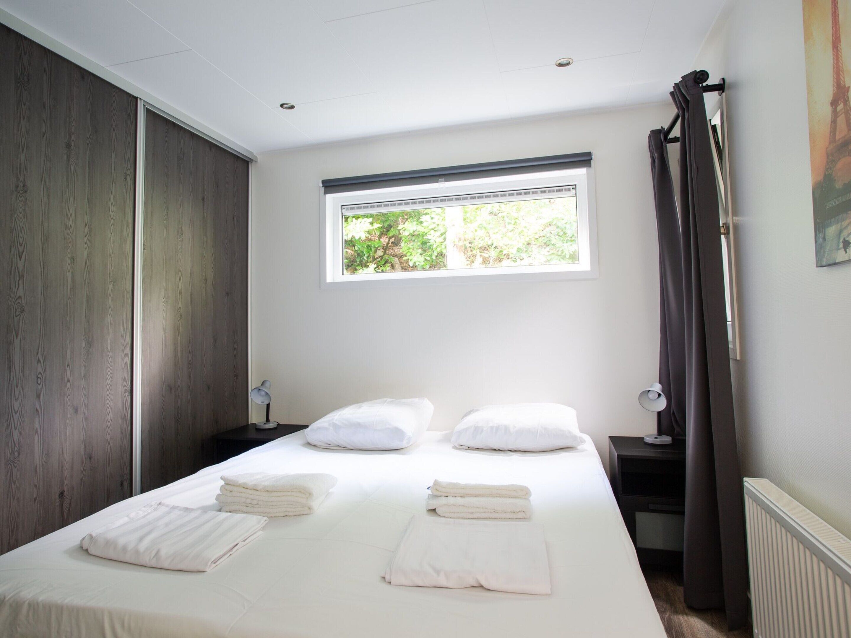 A bedroom with a large bed, a window with a view of greenery, a nightstand with a lamp, and a curtain.