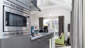 Cocina compacta con microondas, estufa y lavabo integrados. Se ve un sofá verde y una ventana con cortinas al fondo.