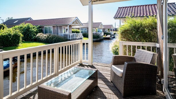 Una terraza cubierta al aire libre con mobiliario de mimbre, una mesa con cubierta de vidrio y vistas a un canal con casas al fondo.