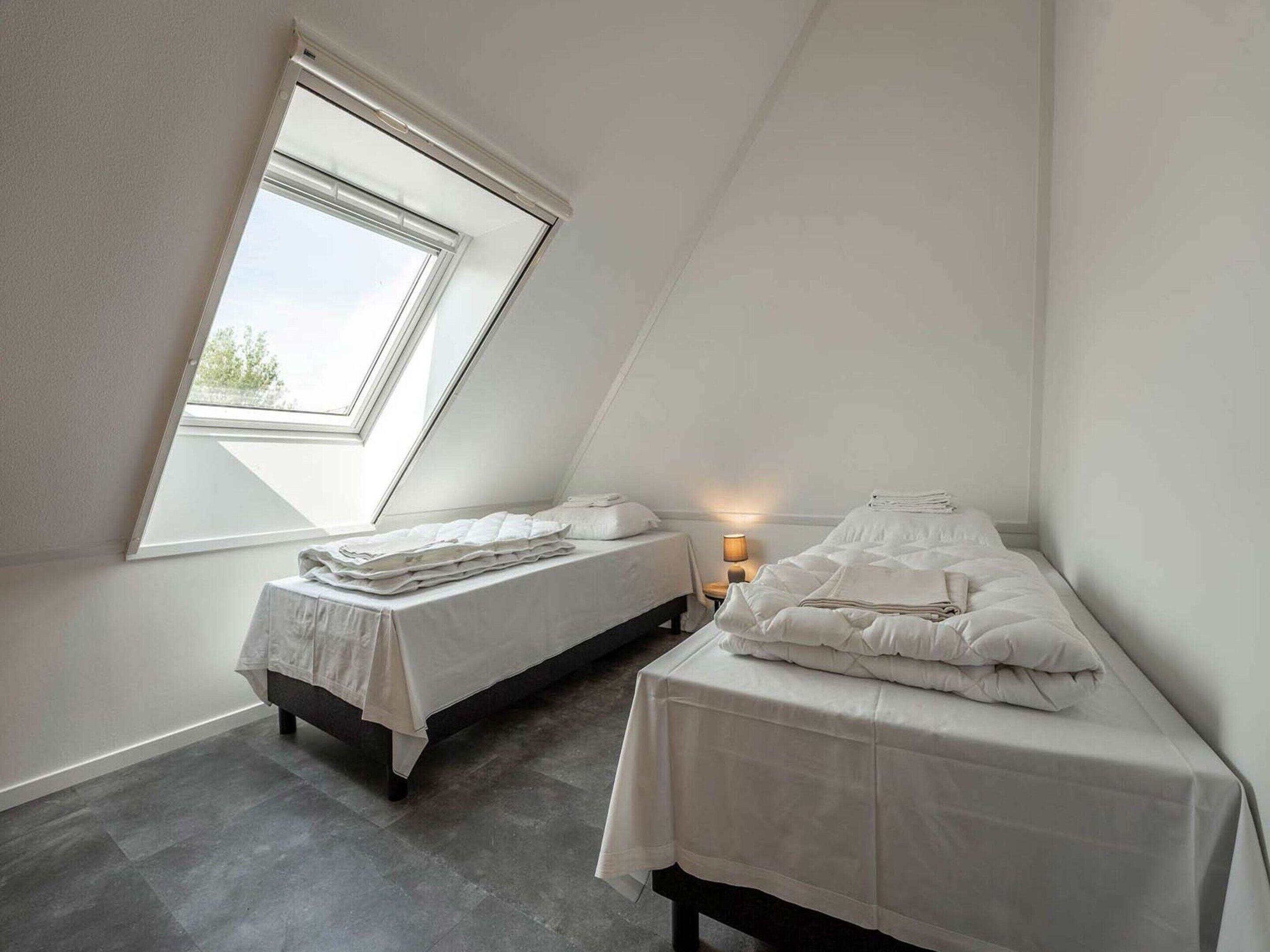 A small, well-lit attic bedroom with two beds, a skylight, and a nightstand with a lamp.