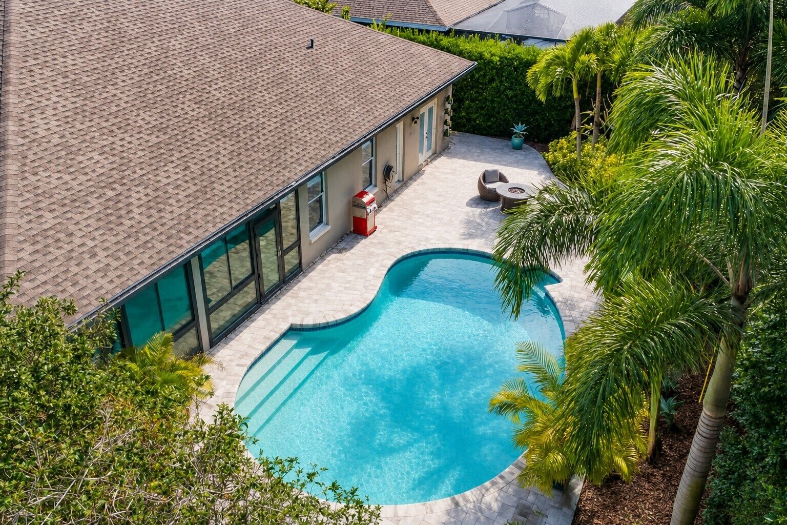 A swimming pool with a curved edge, surrounded by a patio area with outdoor furniture, and a building with large windows and a tiled roof.