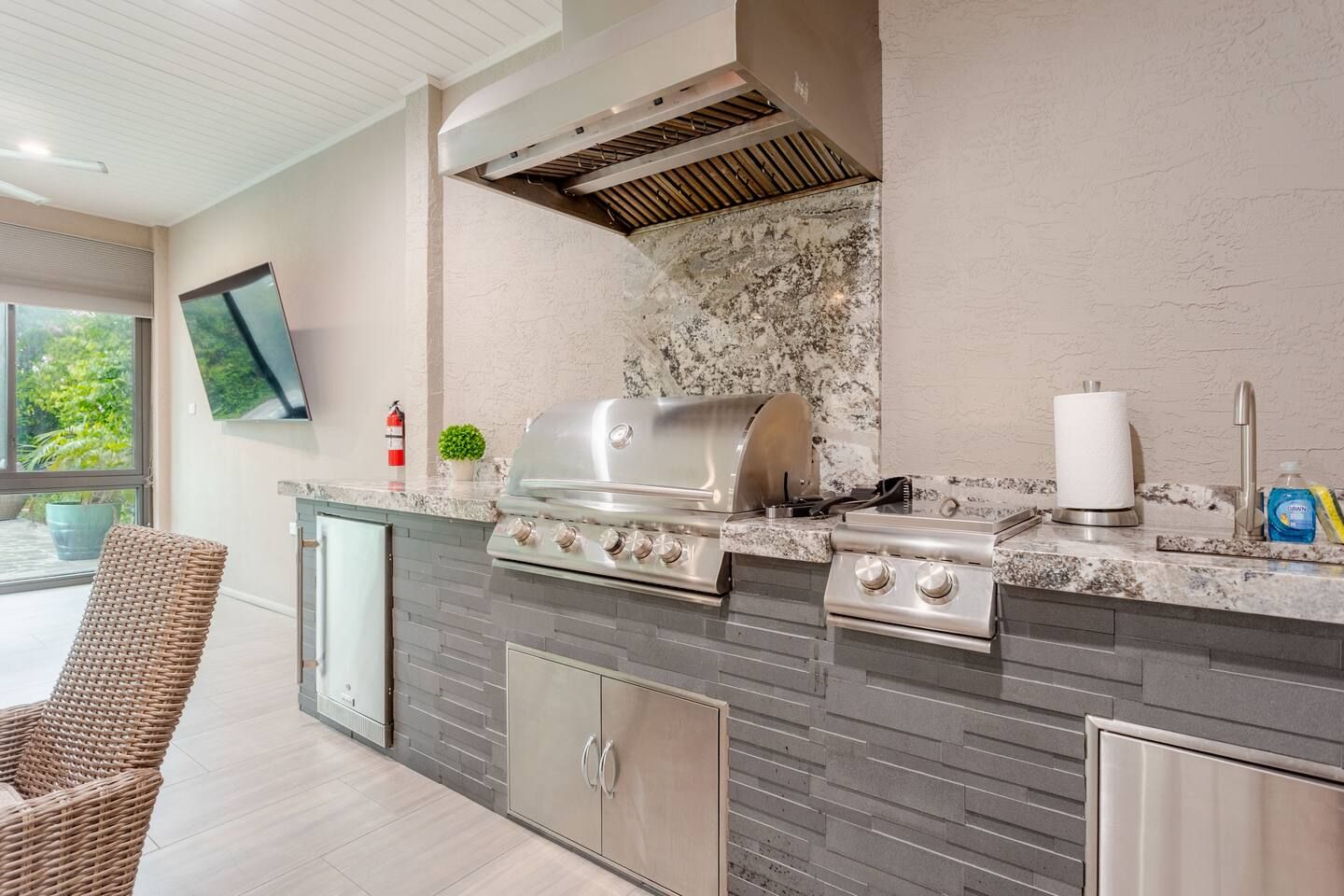 An outdoor kitchen with a grill, a sink, and a television mounted on the wall.