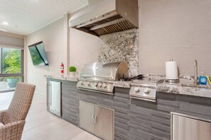 Una cocina exterior con una parrilla, un lavamanos y un televisor montado en la pared.