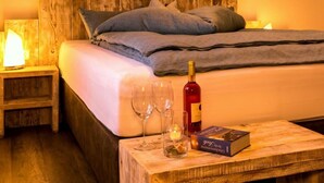 A neatly made bed with pillows, a bedside table with a lamp, two wine glasses, a bottle of wine, and a book on a wooden nightstand. - Dreamtime Modern Retreat (Zinnowitz)