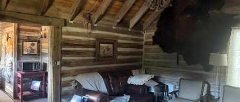A rustic log cabin interior with a leather couch, a white armchair, a wooden coffee table, and a chandelier.