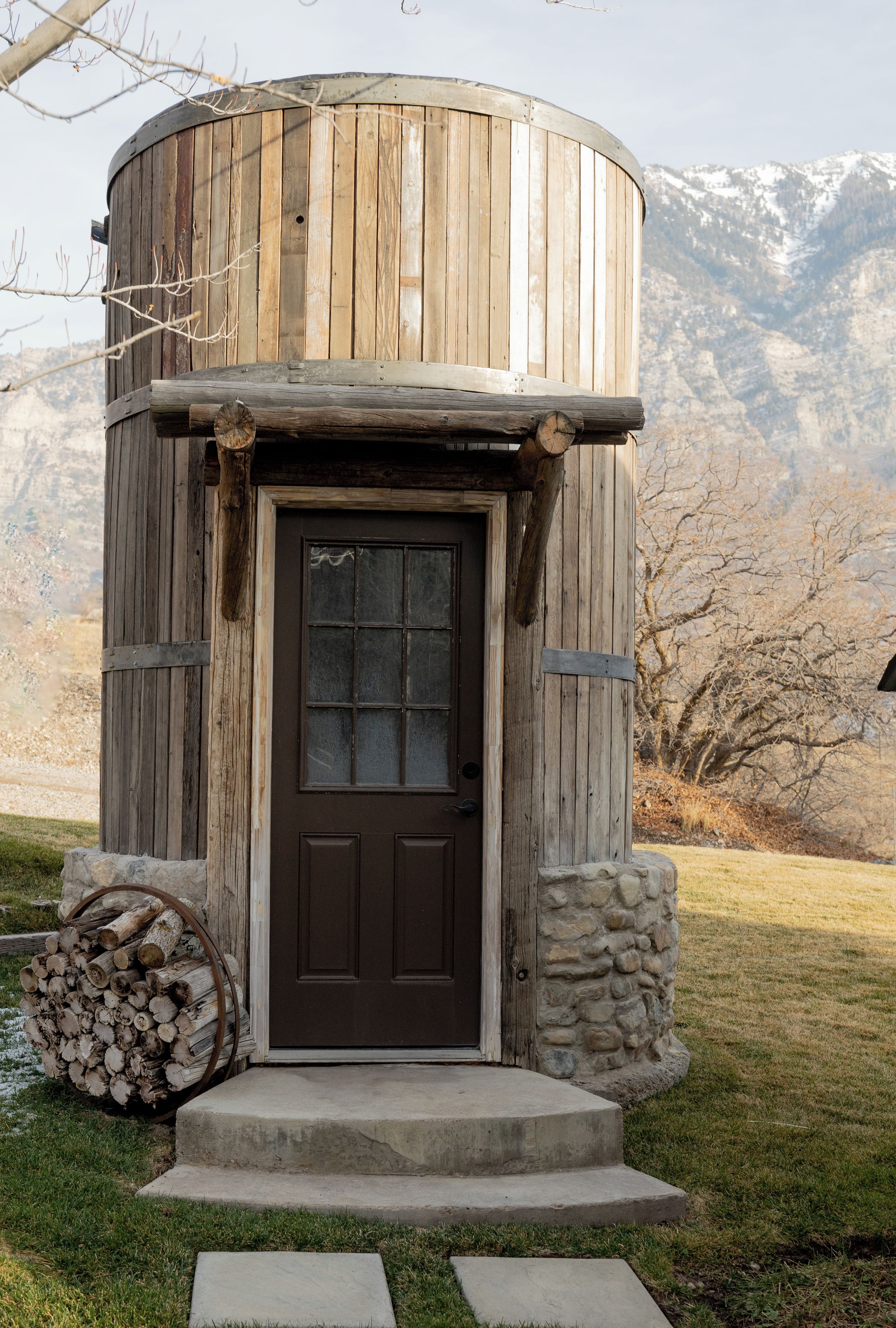 A rustic wooden structure with a dark brown door, a stone base, and a log pile beside it.