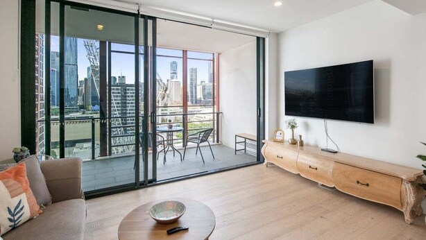 Un soggiorno moderno con un'ampia porta-finestra scorrevole che dà su un balcone, una TV a schermo piatto fissata al muro e un tavolino da caffè in legno.