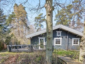 Une cabane en rondins avec un toit à pignon, entourée d’arbres et dotée d’une terrasse en bois.