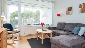 Ein modernes Wohnzimmer mit einem grauen Sofa, einem Holzschaukelstuhl, einem kleinen runden Tisch mit Blumenvase und einem großen Fenster mit Jalousien.