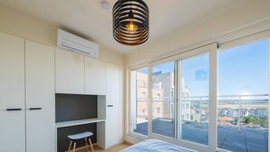 A bedroom with a large bed, a TV on a shelf, a small stool, and a sliding glass door to a balcony with a city view.