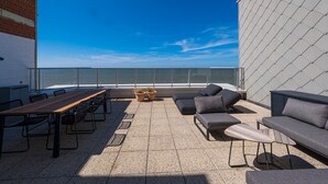 A rooftop terrace with a wooden table, chairs, and a sofa, overlooking a clear blue sky and a distant shoreline.