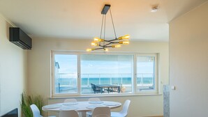 A dining area with a round table, white chairs, and a view of the sea through large windows.
