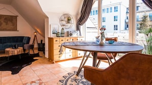 A cozy living room with a dark blue sofa, wooden cabinet, and a dining table with chairs. There is a large window with curtains, a world map on the wall, and a hanging light fixture. - Holiday Apartment in Otseebad Wustrow (Ostseebad Wustrow)