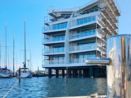 Un edificio de varios pisos con balcones, un puerto deportivo con barcos y un cielo despejado.