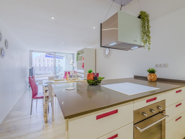 A modern kitchen with a central island, a dining area with pink chairs, and a view of a balcony.