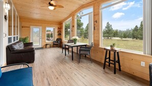 Interior de una cabaña de madera con comedor, sofá y ventanas amplias que dan a una zona boscosa.