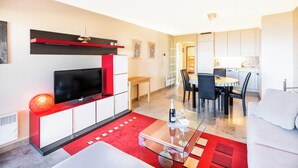 Een moderne woonkamer met een flatscreen televisie, een glazen salontafel en een rood tapijt. Er is een eethoek met een witte tafel en stoelen, en een keuken met witte kasten.