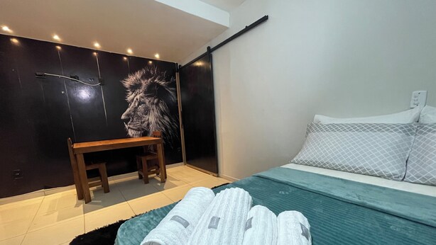 A modern bedroom with a bed, a desk, and a chair. There are towels and a remote control on the bed.
